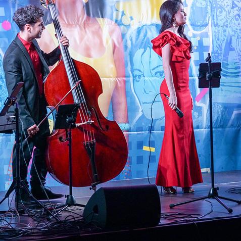 Musikgruppe auf einer Bühne: Pianistin, Kontrabassist und Sängerin in auffälligen Outfits vor buntem, grafischem Hintergrund.