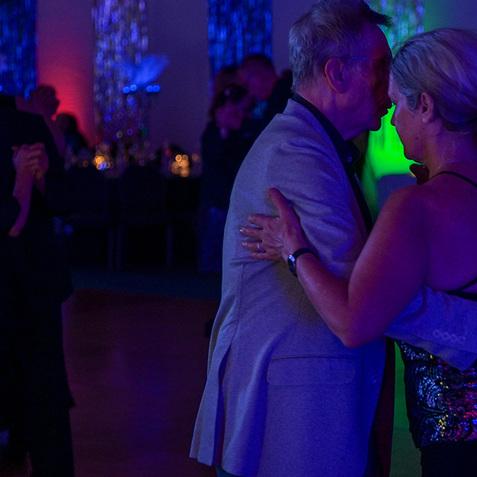 Zwei Tänzer umarmen sich beim Tanzen in einem festlich beleuchteten Saal. Bunte Lichtreflexe im Hintergrund.