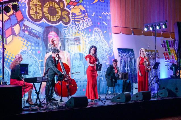 Auftritt einer Musikgruppe in roten Outfits vor einer bunten 80er-Jahre Kulisse mit Mikrofon und Instrumenten.