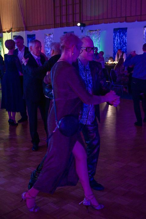 Tanzende Paare in eleganter Kleidung umgeben von bunten Lichtern in einem festlichen Saal.