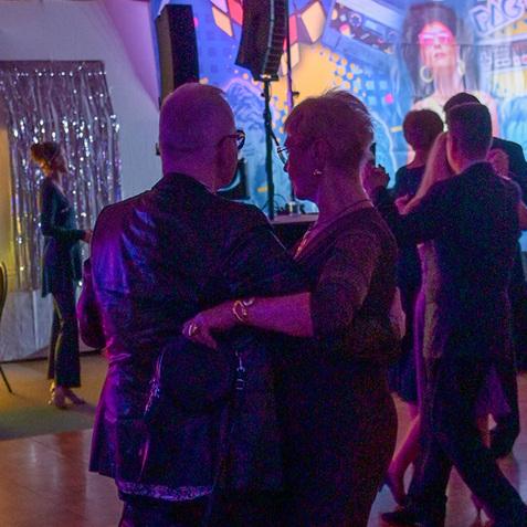 Tanzendes Paar in eleganter Kleidung auf einer festlichen Veranstaltung mit 80er-Jahre-Motiv im Hintergrund.