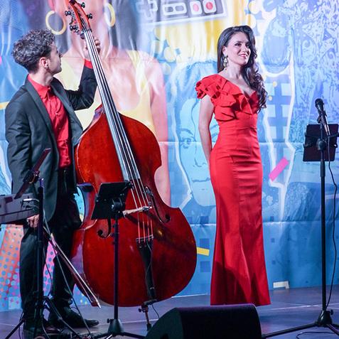 Zwei Musiker auf einer Bühne vor einem bunten 80er-Jahre-Hintergrund, eine Sängerin in rotem Kleid und ein Kontrabassist.