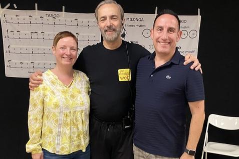 Drei Personen lächeln, stehen im Halbkreis vor einer schwarzen Wand mit Musiknoten und Stühlen im Hintergrund.