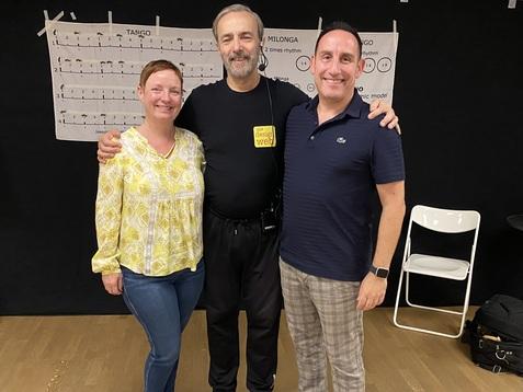 Drei Personen lächeln, stehen im Halbkreis vor einer schwarzen Wand mit Musiknoten und Stühlen im Hintergrund.