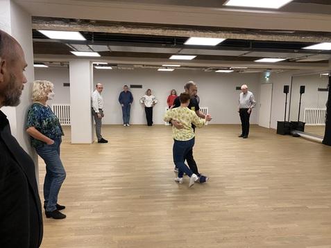 Tanzpaar in einem hellen Raum, umgeben von Zuschauern, während einer Tanzstunde oder Übung.