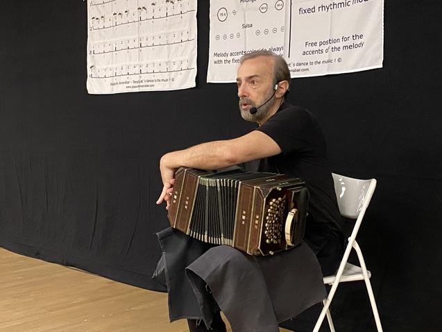 Mann mit Mikrofon sitzt auf einem Stuhl und hält ein Akkordeon, im Hintergrund Lehrmaterial für Tanzrhythmen.