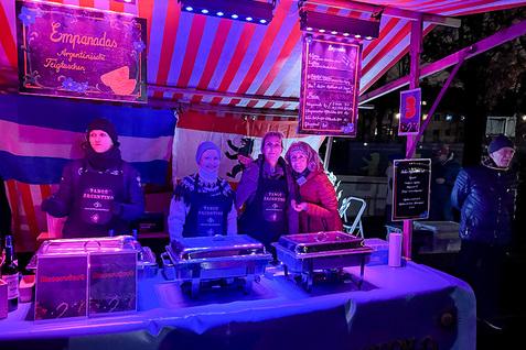 Stand mit argentinischen Empanadas, beleuchtet, mit drei Verkäuferinnen in Schürzen und einer Länderflagge im Hintergrund.