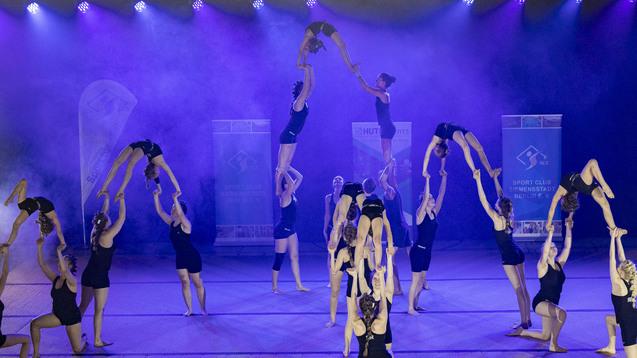 Akrobatik-Darbietung mehrerer Turnerinnen auf einer Bühne mit lila Beleuchtung und Banner im Hintergrund.