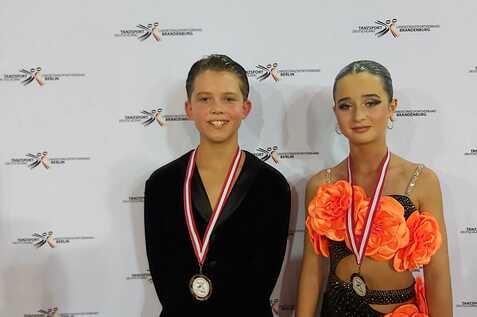 Zwei T&auml;nzer stehen vor einem Banner der Landesmeisterschaften in Berlin, beide tragen Medaillen und festliche Kleidung.
