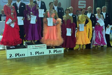 Siegerteam der Tanzmeisterschaft in Berlin, auf dem Treppchen mit Urkunden und Medaillen in festlicher Kleidung.