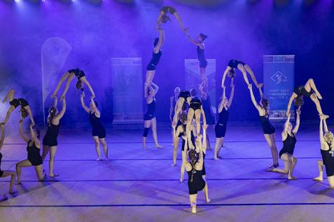 Akrobatik-Darbietung mehrerer Turnerinnen auf einer B&uuml;hne mit lila Beleuchtung und Banner im Hintergrund.