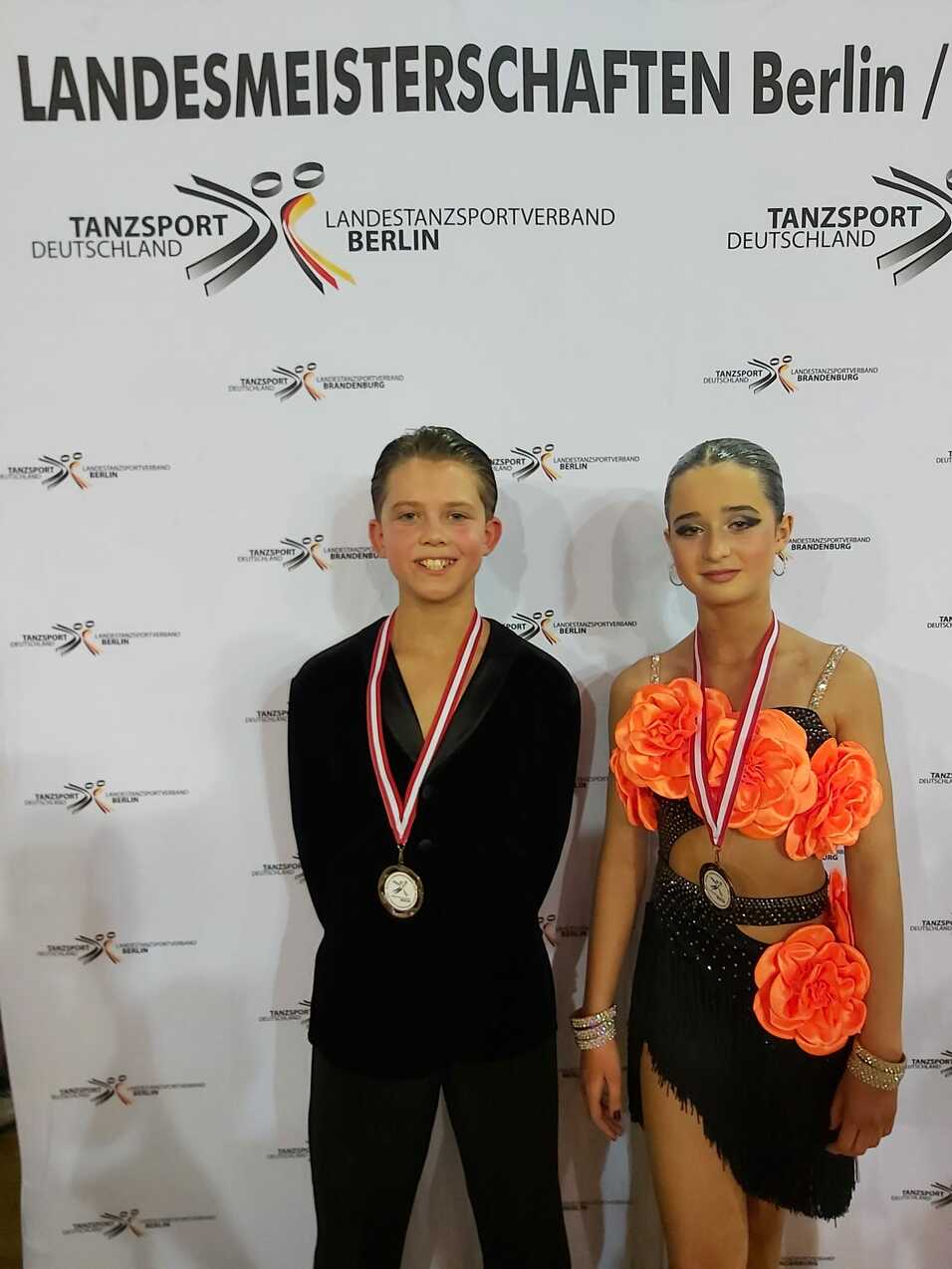 Zwei Tänzer stehen vor einem Banner der Landesmeisterschaften in Berlin, beide tragen Medaillen und festliche Kleidung.