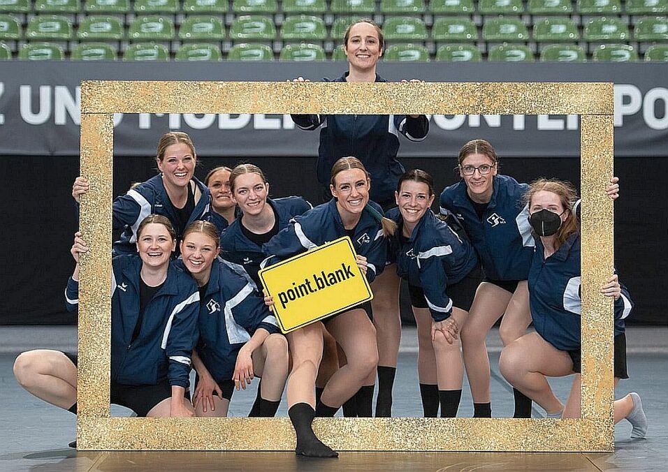 Gruppe von Tänzerinnen in blauen Trainingsanzügen posiert mit einem goldenen Bilderrahmen und einem Schild mit der Aufschrift "point.blank".