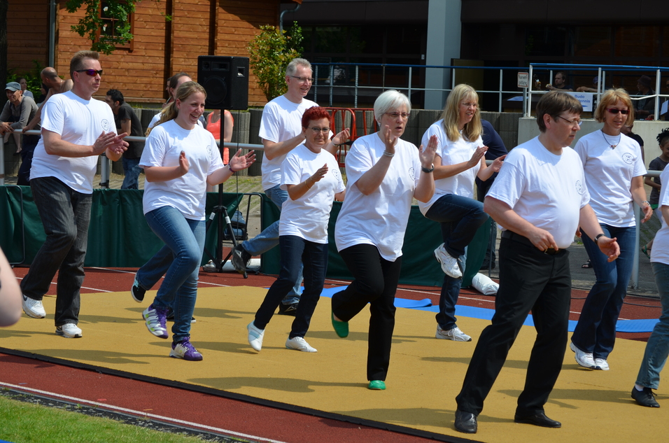 Gruppe von Menschen in weißen T-Shirts, die zusammen auf einer Sportfläche tanzen und klatschen.