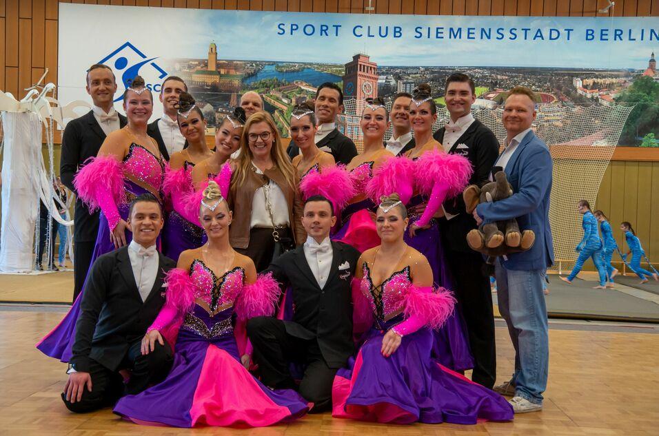 Gruppenfoto von Tänzern in bunten Kostümen mit Federärmel und Glitzer, posierend in einer Sporthalle. Hintergrund mit Banner.