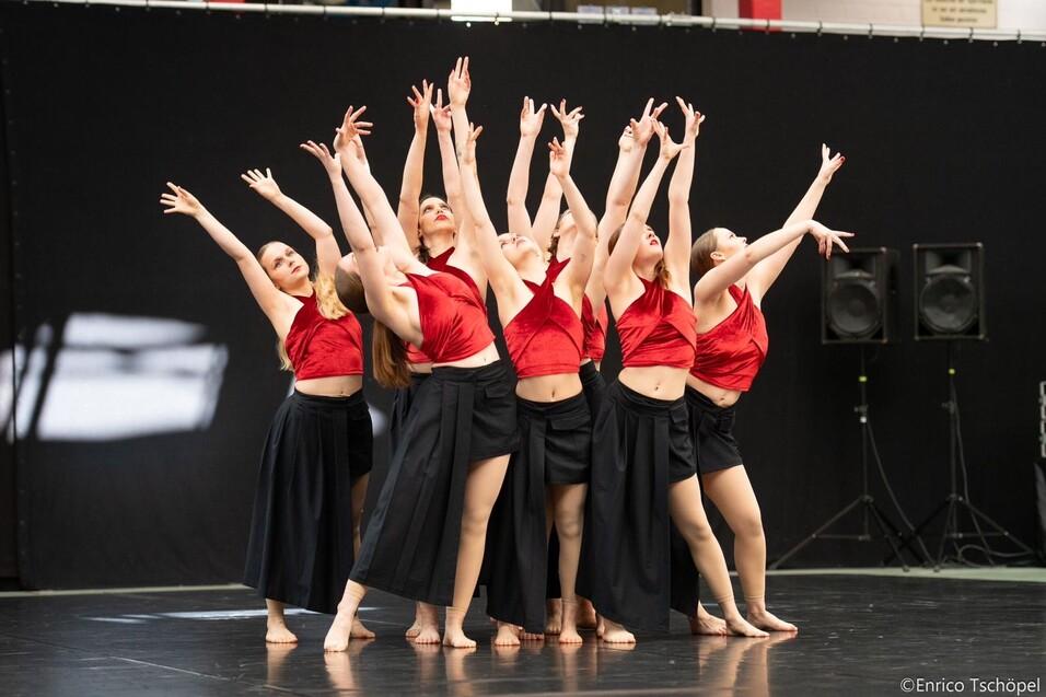 Gruppe von Tänzerinnen in roten Oberteilen und schwarzen Röcken, die synchron auf einer Bühne tanzen.