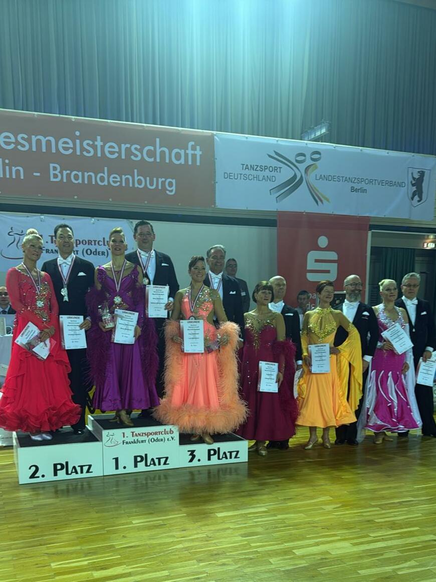 Siegerteam der Tanzmeisterschaft in Berlin, auf dem Treppchen mit Urkunden und Medaillen in festlicher Kleidung.