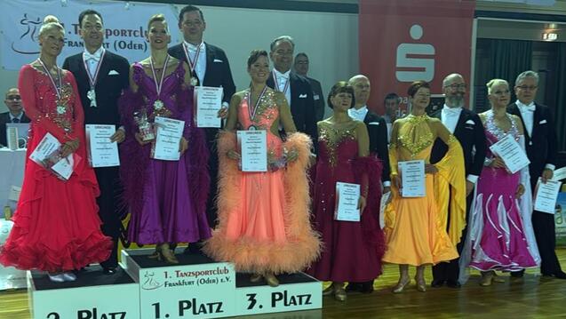 Siegerteam der Tanzmeisterschaft in Berlin, auf dem Treppchen mit Urkunden und Medaillen in festlicher Kleidung.