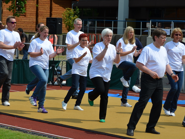 Gruppe von Menschen in wei&szlig;en T-Shirts, die zusammen auf einer Sportfl&auml;che tanzen und klatschen.