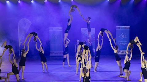 Akrobatik-Darbietung mehrerer Turnerinnen auf einer Bühne mit lila Beleuchtung und Banner im Hintergrund.