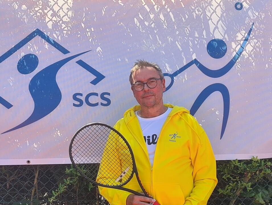 Mann in gelber Jacke mit Tennisschl&auml;ger vor einem Banner mit Logo des SCS und einer spielenden Figur.