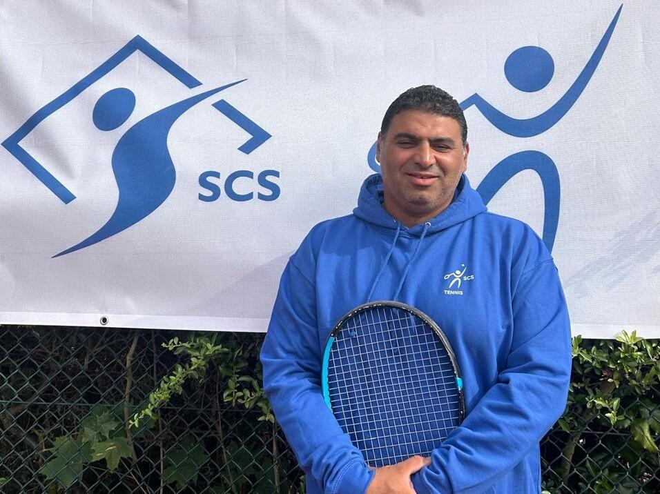 Mann in blauer Jacke h&auml;lt einen Tennisschl&auml;ger, steht vor einem Banner mit dem Logo von SCS.