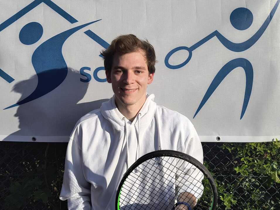 Junger Mann in weißem Hoodie hält einen Tennisschläger vor einem Banner mit Sportmotiven.