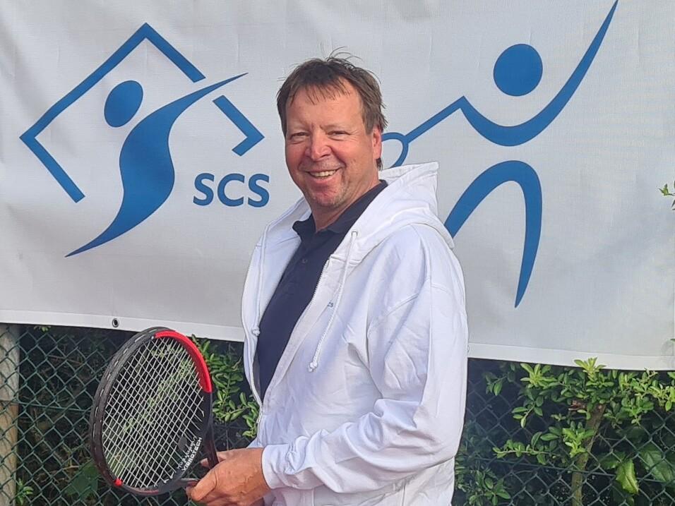 L&auml;chelnder Mann mit Tennisschl&auml;ger in der Hand steht vor einem Banner mit dem Logo der SCS-Tennisgemeinschaft.