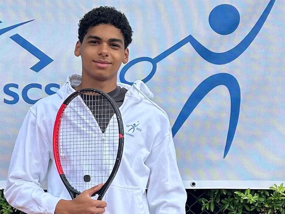 Junger Tennis-Spieler hält einen Tennisschläger in der Hand vor einem Banner mit Tennis-Motiv.