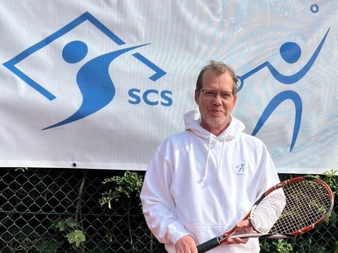 Mann mit Brille in einem wei&szlig;en Hoodie h&auml;lt einen Tennisschl&auml;ger vor einem SCS-Logo an einem Tennisplatz.