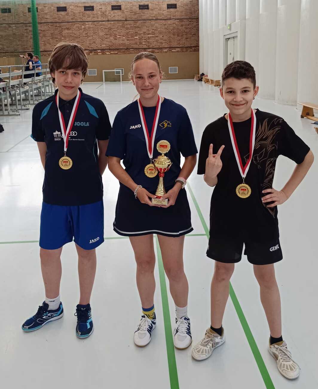 Drei Tischtennisspieler stehen in einer Sporthalle, jeder trägt eine Medaille, der mittlere hält einen Pokal.