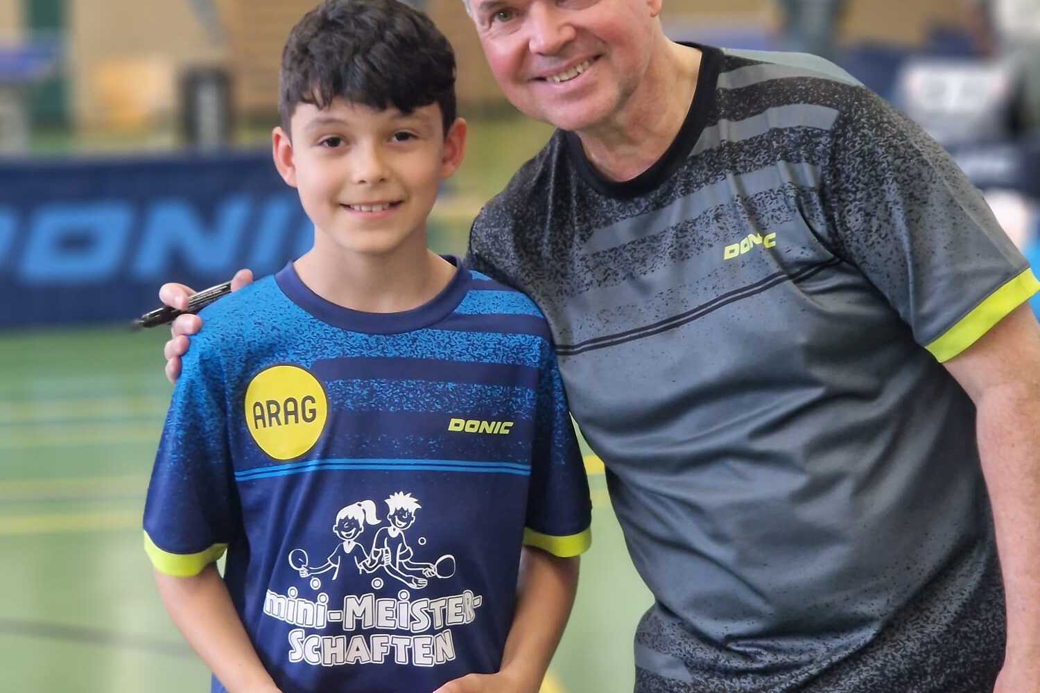 Portr&auml;t eines Jungen mit Tischtennisschl&auml;ger und einem Mann in Sportkleidung, Hintergrund: Tischtennishalle.