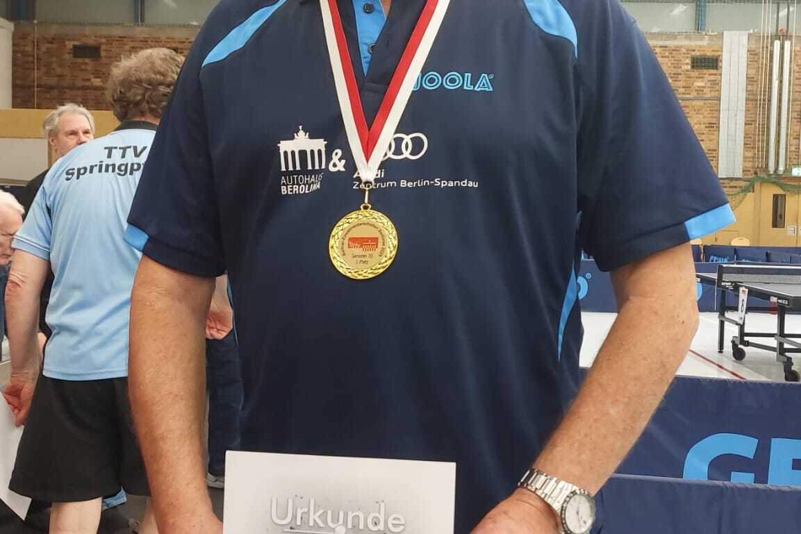 Mann mit Brille hält eine Urkunde und Medaille nach Tischtennisturnier, steht in einer Sporthalle.