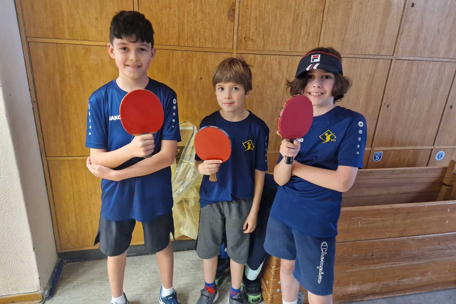 Drei Kinder in blauen Sportshirts halten Tischtennisschläger vor einer Holzwand und einer Bank.