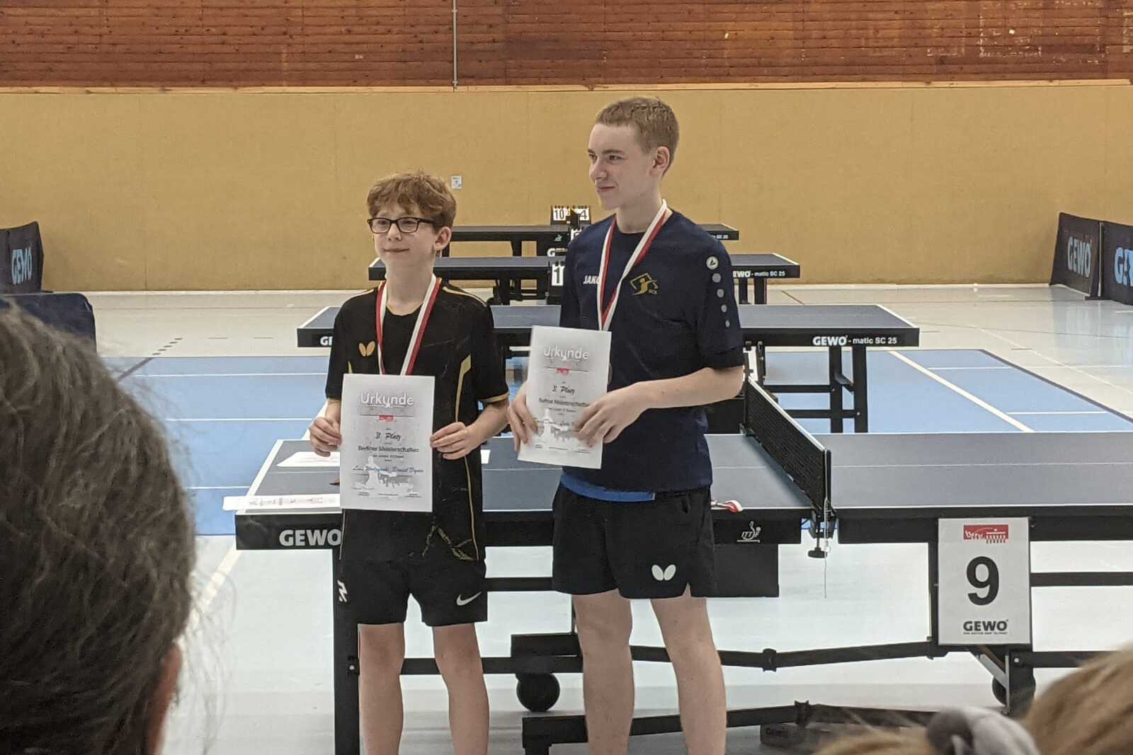 Zwei Tischtennisspieler stehen mit Urkunden und Medaillen auf einem Tischtennisturnier, Tischtennisplatten im Hintergrund.