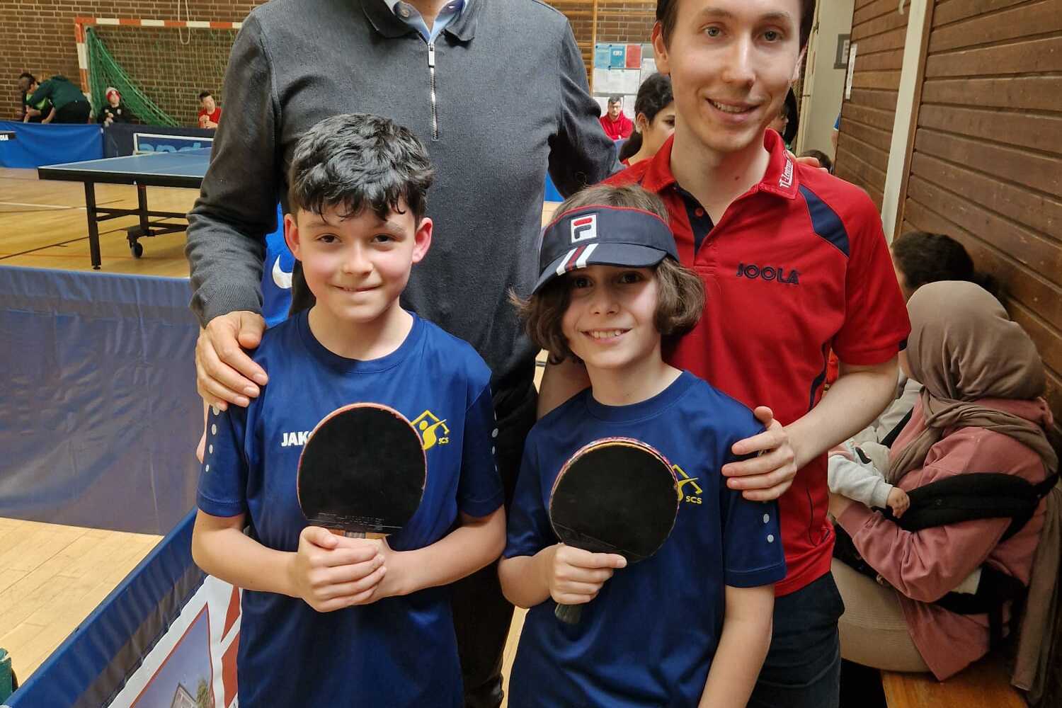 Vier Personen stehen in einer Sporthalle zusammen, zwei Kinder mit Tischtennisschlägern und zwei Erwachsene im Hintergrund.