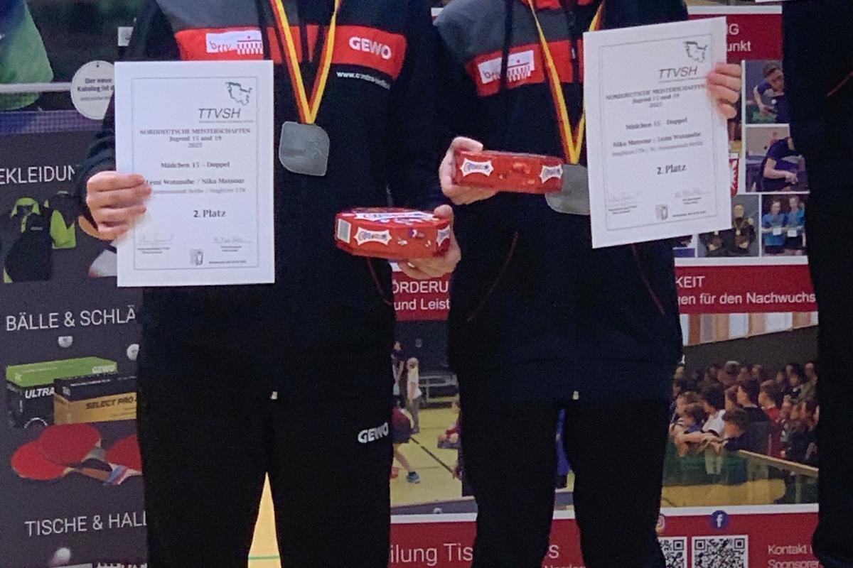 Zwei Mädchen stehen auf dem Siegertreppchen und halten Urkunden für den 1. und 2. Platz in einem Tischtennisturnier.