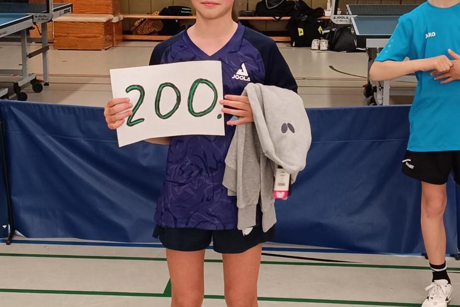 Mädchen hält ein Plakat mit der Zahl „200“ in einer Sporthalle, während ein weiterer Junge im Hintergrund steht.