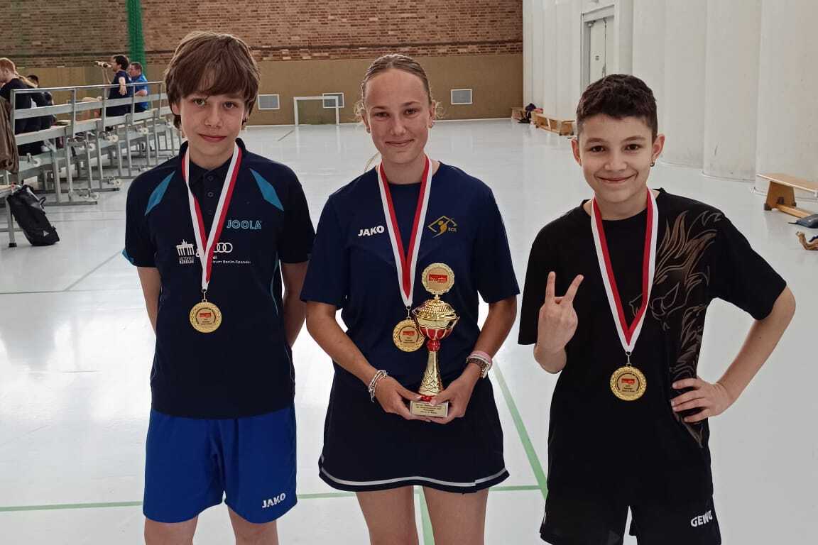 Drei Tischtennisspieler stehen in einer Sporthalle, jeder trägt eine Medaille, der mittlere hält einen Pokal.