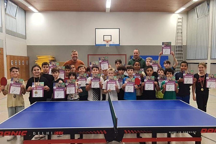 Kinder stehen in einer Sporthalle mit Urkunden und Medaillen vor einem Tischtennistisch.