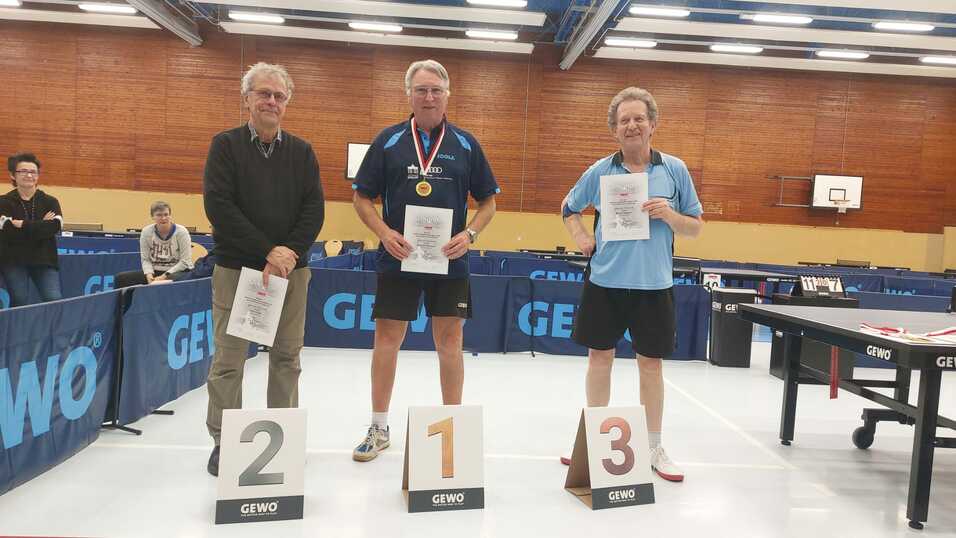 Drei Männer in Sportkleidung stehen mit Urkunden und Medaillen vor einem Tischtennis-Tisch. Anzeigetafeln zeigen die Plätze 1, 2 und 3.