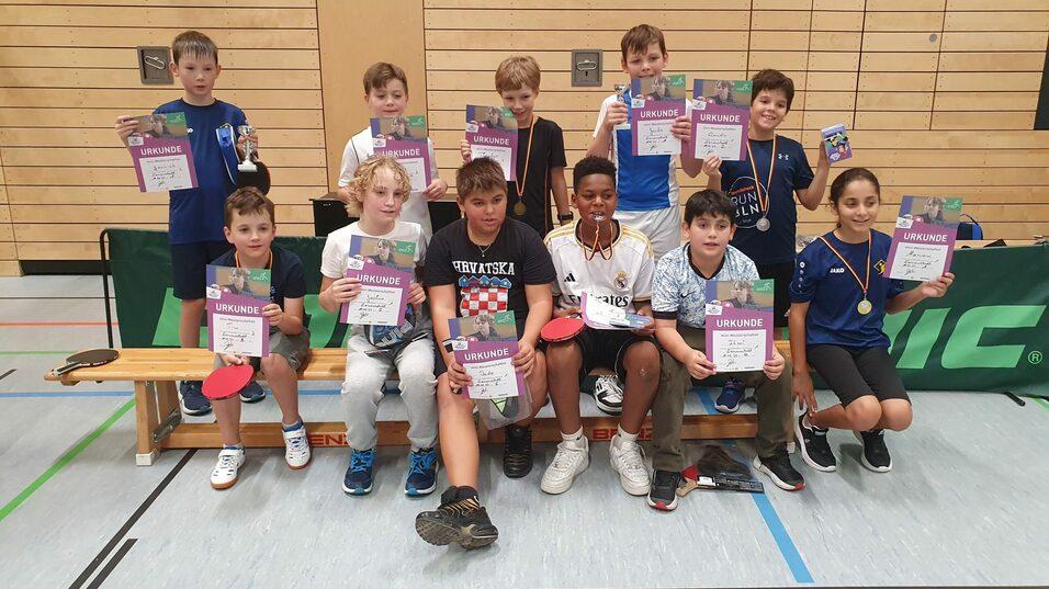 Gruppe von Kindern mit Urkunden und Medaillen nach einem Tischtennisturnier in einer Sporthalle.