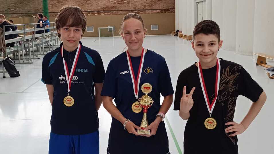 Drei Tischtennisspieler stehen in einer Sporthalle, jeder trägt eine Medaille, der mittlere hält einen Pokal.