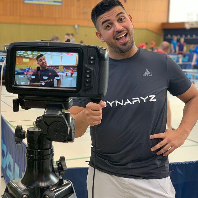 Mann mit T-Shirt steht vor einer Kamera in einem Tischtennis-Hallenambiente und l&auml;chelt in die Linse.