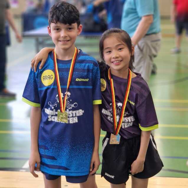 Zwei Kinder in Tischtennis-Trikots stehen l&auml;chelnd, Medaillen um den Hals, in einer Sporthalle.