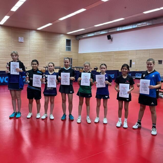 Acht Mädchen in Sportkleidung halten Siegerurkunden und Pokale in einer Tischtennishalle mit roten Böden.