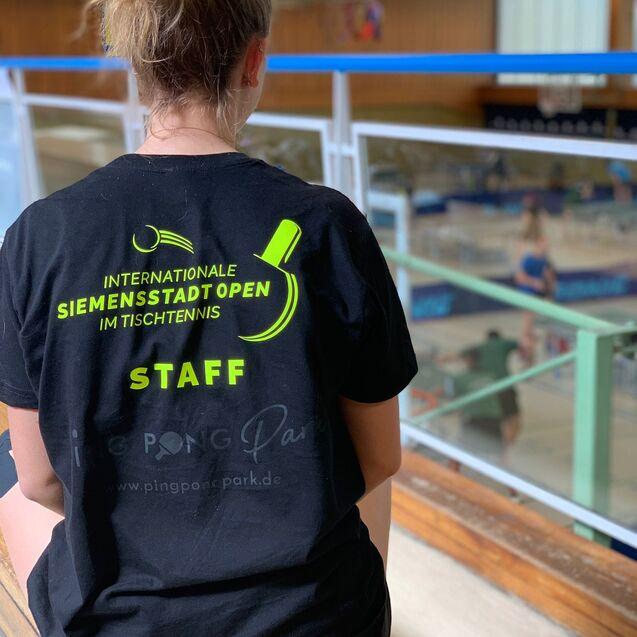 R&uuml;ckansicht einer Person in schwarzem Shirt mit Aufdruck &bdquo;Internationale Siemenstadt Open im Tischtennis STAFF&ldquo;.