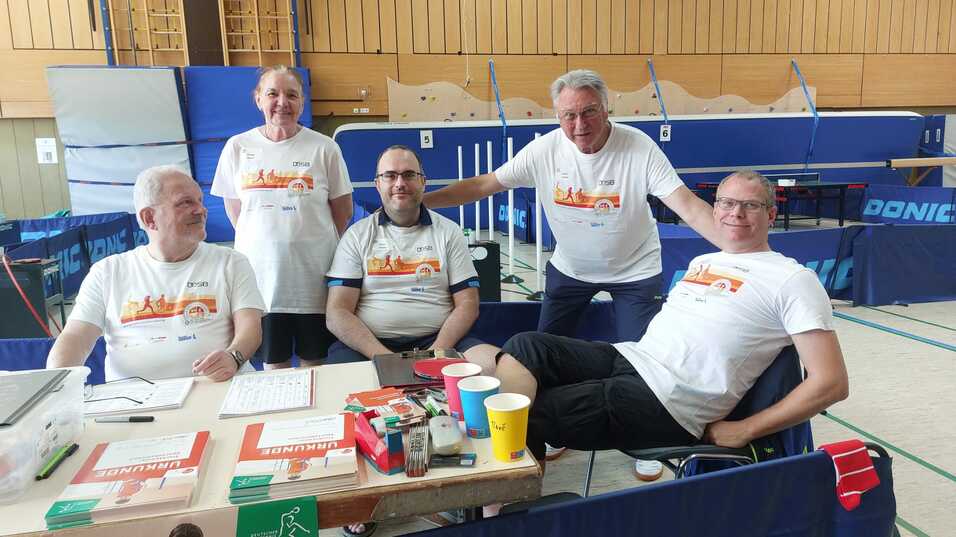 Fünf Personen in einheitlichen Shirts sitzen an einem Tisch mit Getränken und Unterlagen in einer Sporthalle.
