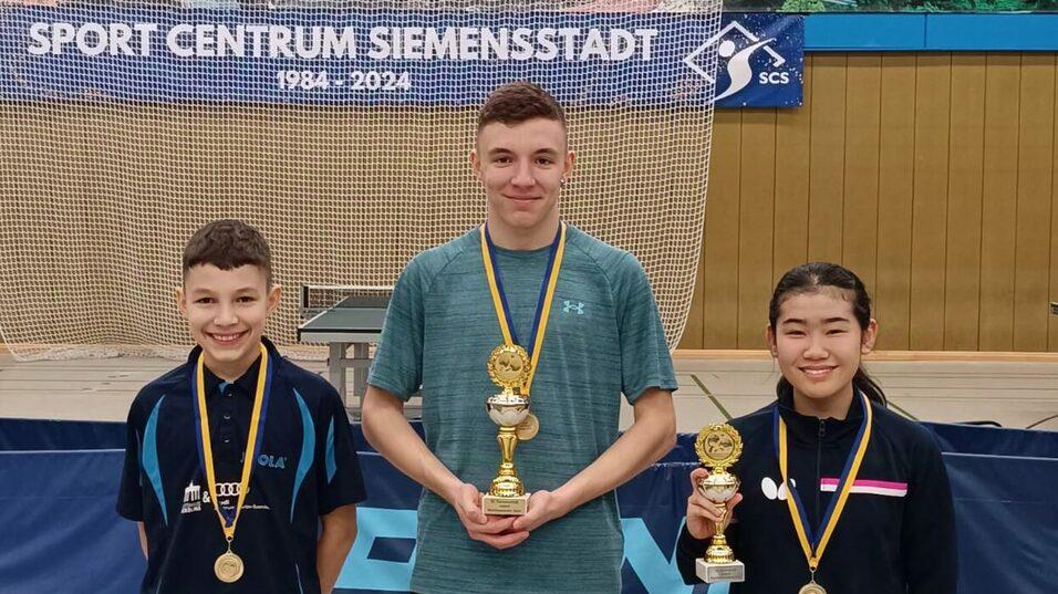 Drei Tischtennisspieler stehen mit Pokalen und Medaillen in einer Sporthalle, im Hintergrund ein Netz und Banner.