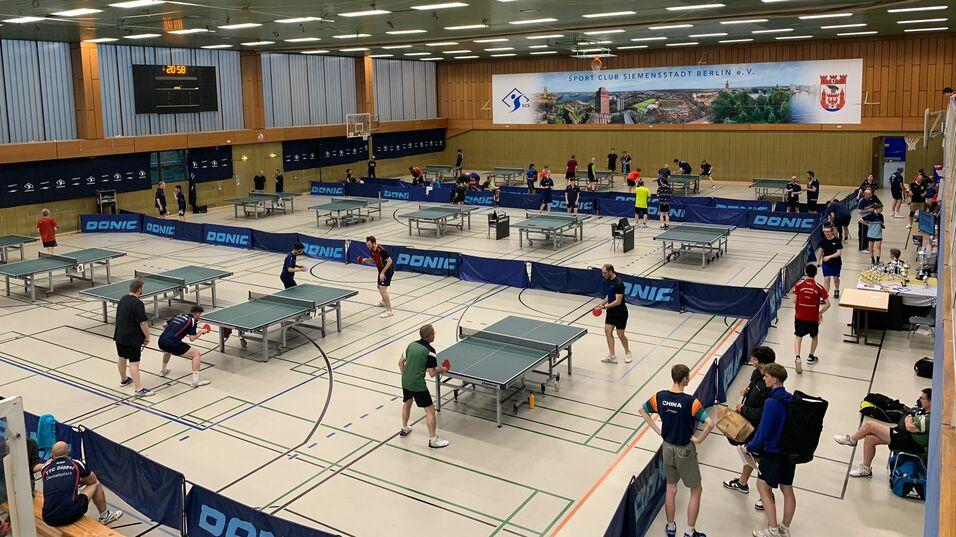 Tischtennisveranstaltung in einer Sporthalle mit mehreren Spielern an Tischen und einer digitalen Anzeigetafel im Hintergrund.